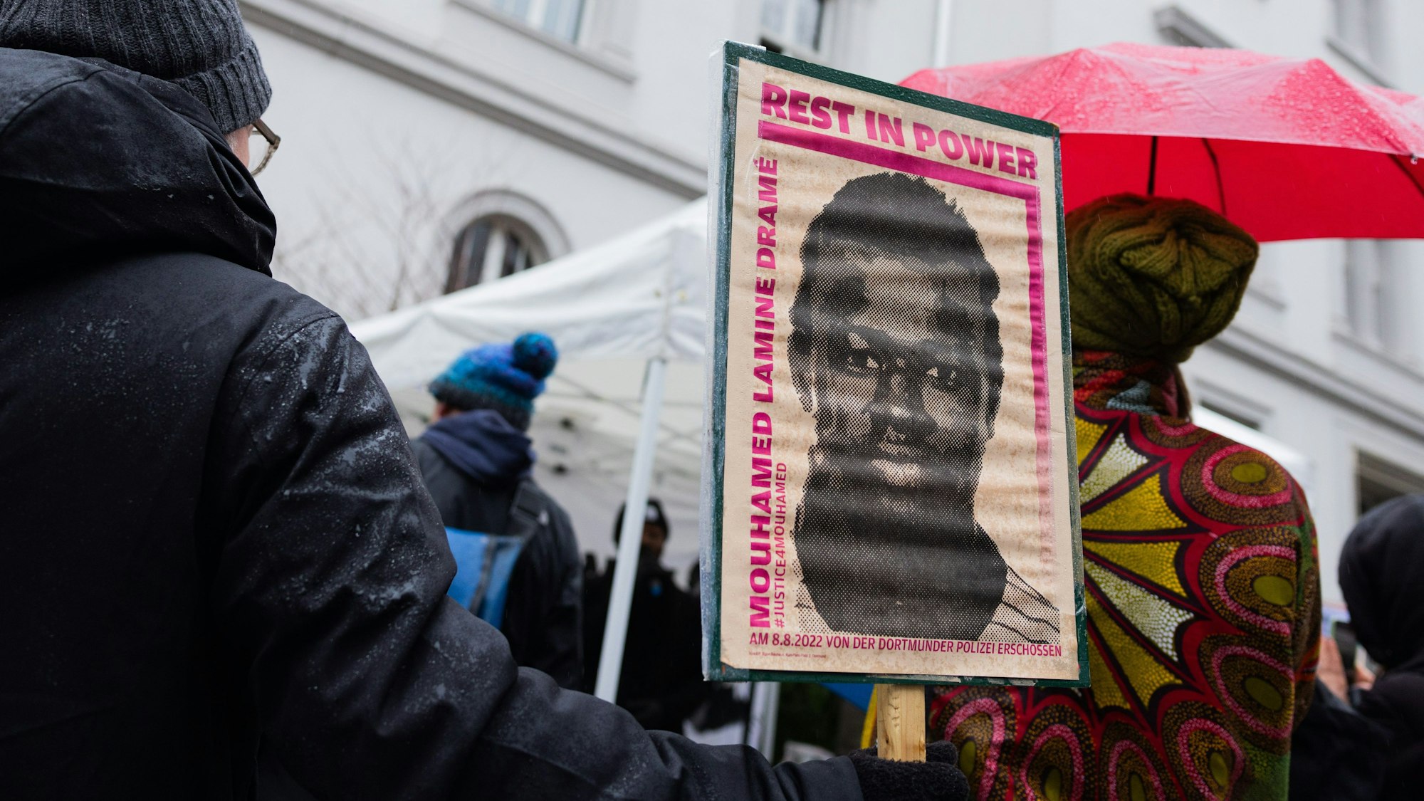 PRODUKTION - 19.12.2023, Nordrhein-Westfalen, Dortmund: Demonstranten stehen mit Plakaten, die das Portrait des 16-jährigen Senegalesen Mouhamed Drame zeigen, vor dem Landgericht. Hier beginnt der Prozess nach tödlichen Polizeischüssen auf den jungen Flüchtling. Der Jugendliche aus dem Senegal war am 8. August 2022 auf dem Gelände einer Jugendhilfeeinrichtung erschossen worden. Foto: Rolf Vennenbernd/dpa +++ dpa-Bildfunk +++