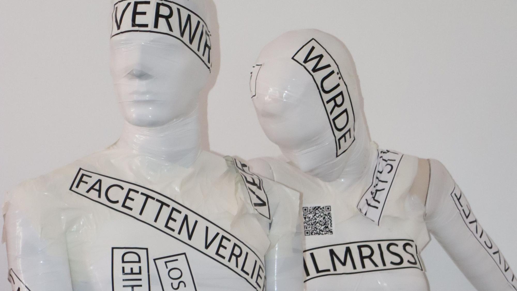 Zu sehen sind zwei weiße Schaufensterpuppen, die mit Worten beklebt sind.