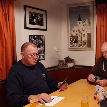 Drei Männer sitzen in einer Kneipe am Tisch und spielen Karten. An der Wand hängt ein Ölbild von E.O. Primbsch, das die Kaller Pfarrkirche zeigt.