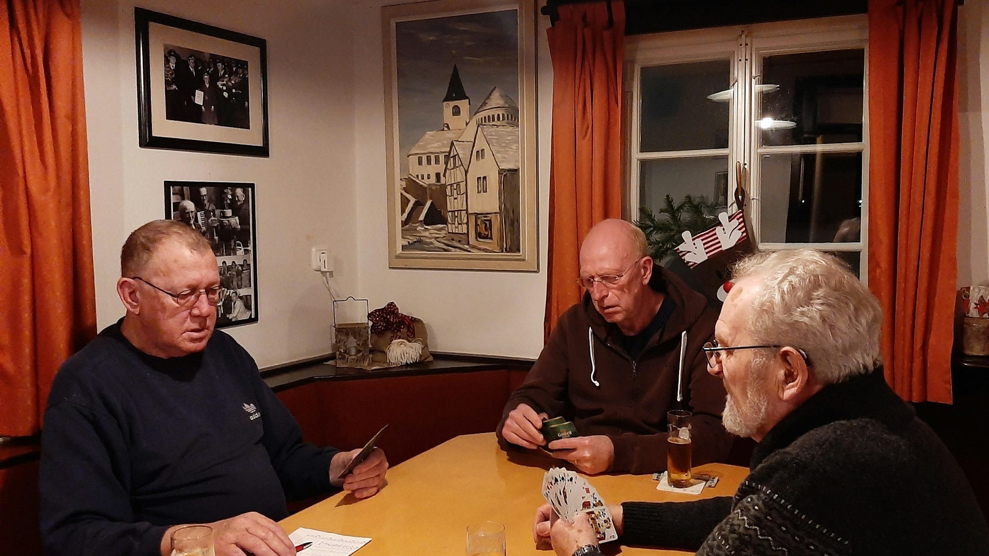Drei Männer sitzen in einer Kneipe am Tisch und spielen Karten. An der Wand hängt ein Ölbild von E.O. Primbsch, das die Kaller Pfarrkirche zeigt.