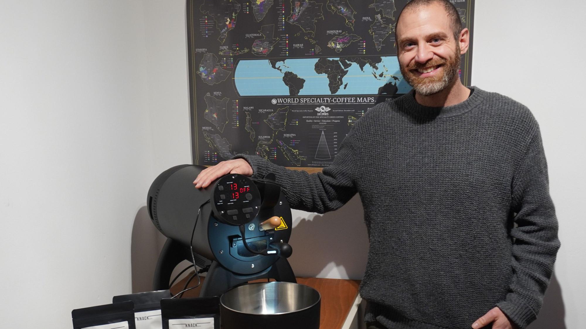 Ein Mann lächelt in die Kamera. Neben ihm auf einem Tisch steht eine Kaffeeröstmaschine.