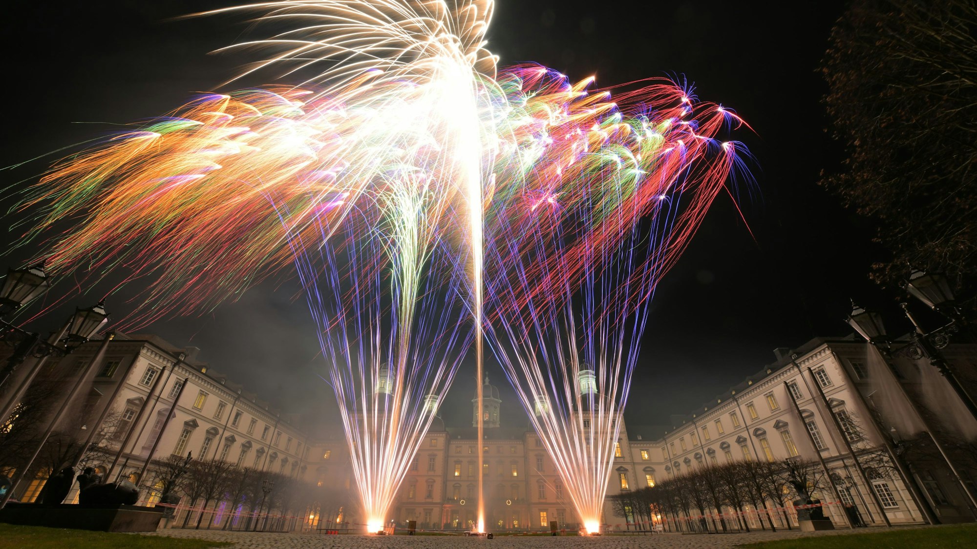 Die Lichter von bunte Feuerwerksraketen sind vor einer Häuserkulisse zu sehen.