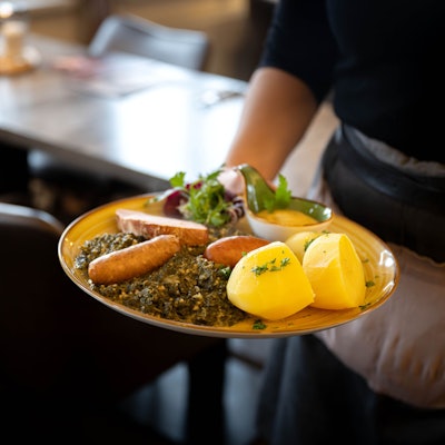 Man sieht den Arm einer Kellnerin, die einen Teller mit Möhren, Kartoffeln, Gehackten und Salat transportiert.