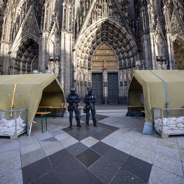Polizisten stehen mit Maschinenpistolen vor dem Kölner Dom. Neben ihnen stehen Zelte.