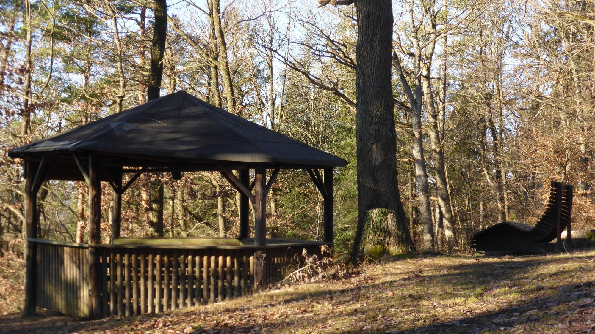 Vor einer Baumreihe sind links eine Wanderhütte und rechts ein hölzerner Liegestuhl zu erkennen.