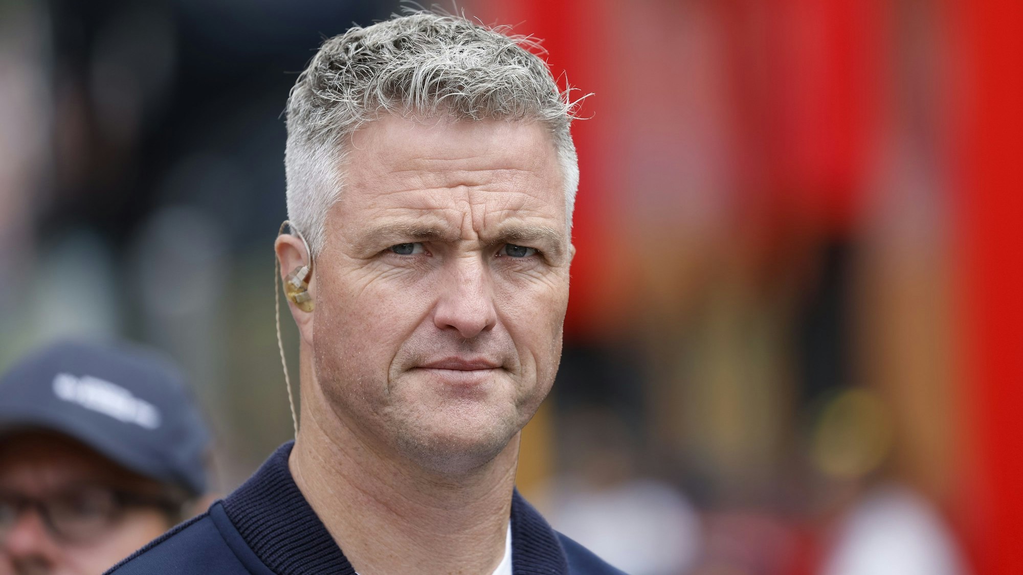 Ralf Schumacher, ehemaliger Formel-1-Pilot aus Deutschland, steht an einer Rennstrecke. Er trägt eine blaue Jacke und ein weißes T-Shirt.