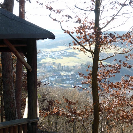 Im Vordergrund ist eine Waldhütte zu sehen. Im Hintergrund erstreckt sich ein mit Häusern gesprenkeltes Tal.