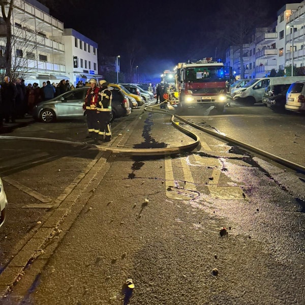 Ein Feuerwehrfahrzeug steht in der Dunkelheit auf einer Straße zwischen Mehrfamilienhäusern, Rettungskräfte und eine Menschengruppe sind zu sehen.