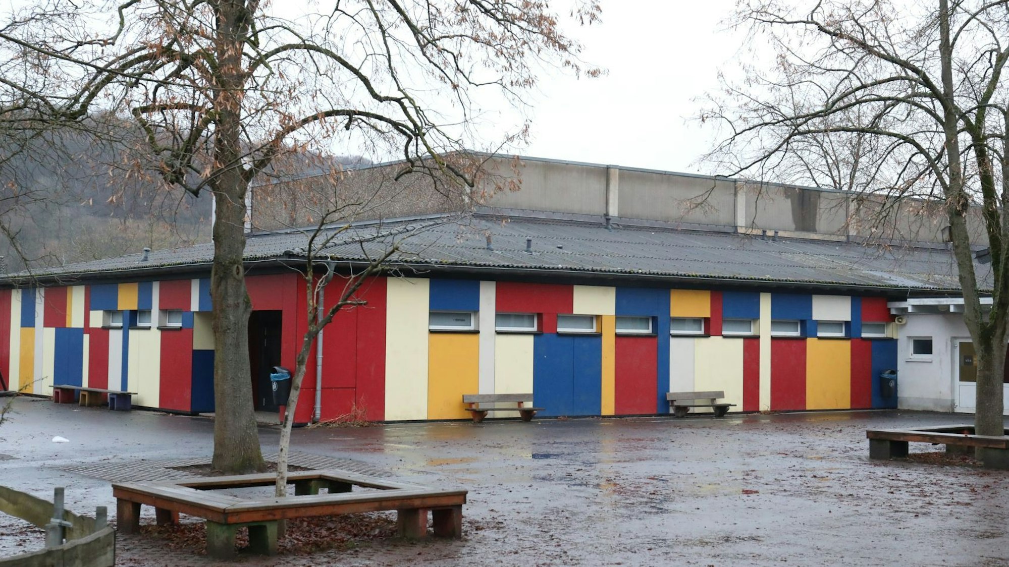 Vor einem flachen Gebäude mit einer bunten Fassade stehen zwei Bäume und einige Sitzbänke.