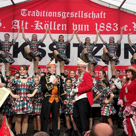 Die Volkssitzung 2023 der KG Alt-Köllen im Zelt auf dem Neumarkt: Präsident der KG spricht mit Festkomitee-Chef Christof Kuckelkorn, im Hintergrund hebt das Tanzkorps Mariechen in die Höhe.