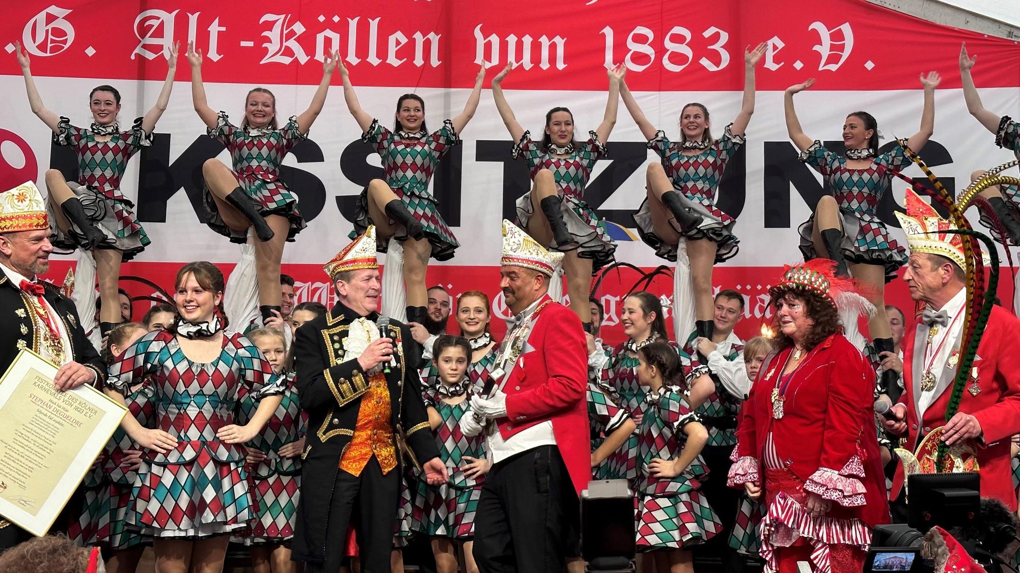 Die Volkssitzung 2023 der KG Alt-Köllen im Zelt auf dem Neumarkt: Präsident der KG spricht mit Festkomitee-Chef Christof Kuckelkorn, im Hintergrund hebt das Tanzkorps Mariechen in die Höhe.