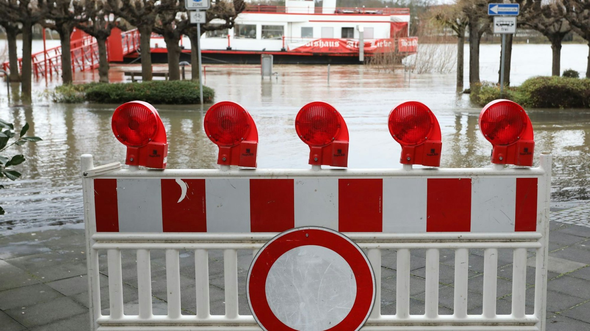 Ein „Durchfahrt verboten“-Schild und eine Absperrung stehen auf der teilweise überfluteten Rheinallee, im Hintergrund ist das Schiff „Alte Liebe“ zu erkennen.
