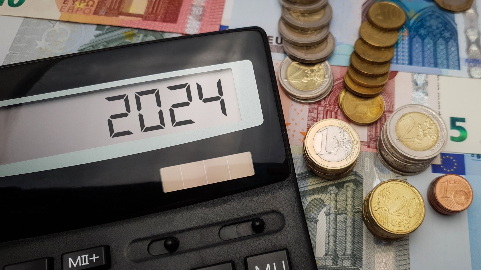 Ein Taschenrechner, auf dem 2024 steht, liegt auf verschiedenen Geldscheinen und -münzen
