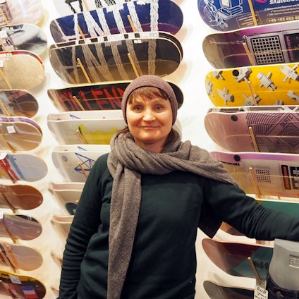 Eine Frau in Pulli, Schal und Mütze steht vor einer Wand, an der Skateboards in vielen Designs hängen.