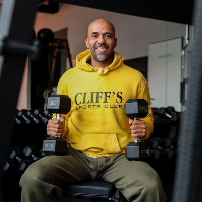 Clifford Opoku-Afari sitzt mit Hanteln in der Hand in einem Sportstudio.
