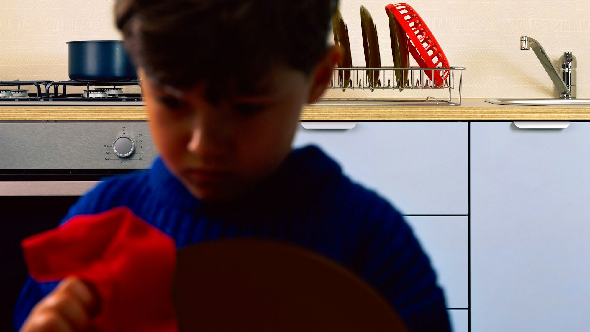 Ein Junge sitzt in einer Küche, im Hintergrund sind ein Waschbecken und Geschirr zu sehen.