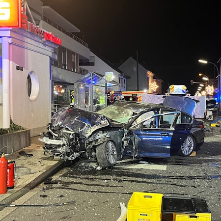 Ein zertrümmertes, dunkles Auto steht quer auf einer Straße, im Hintergrund ist das verbogene Gestänge des Wartehäuschens zu sehen.