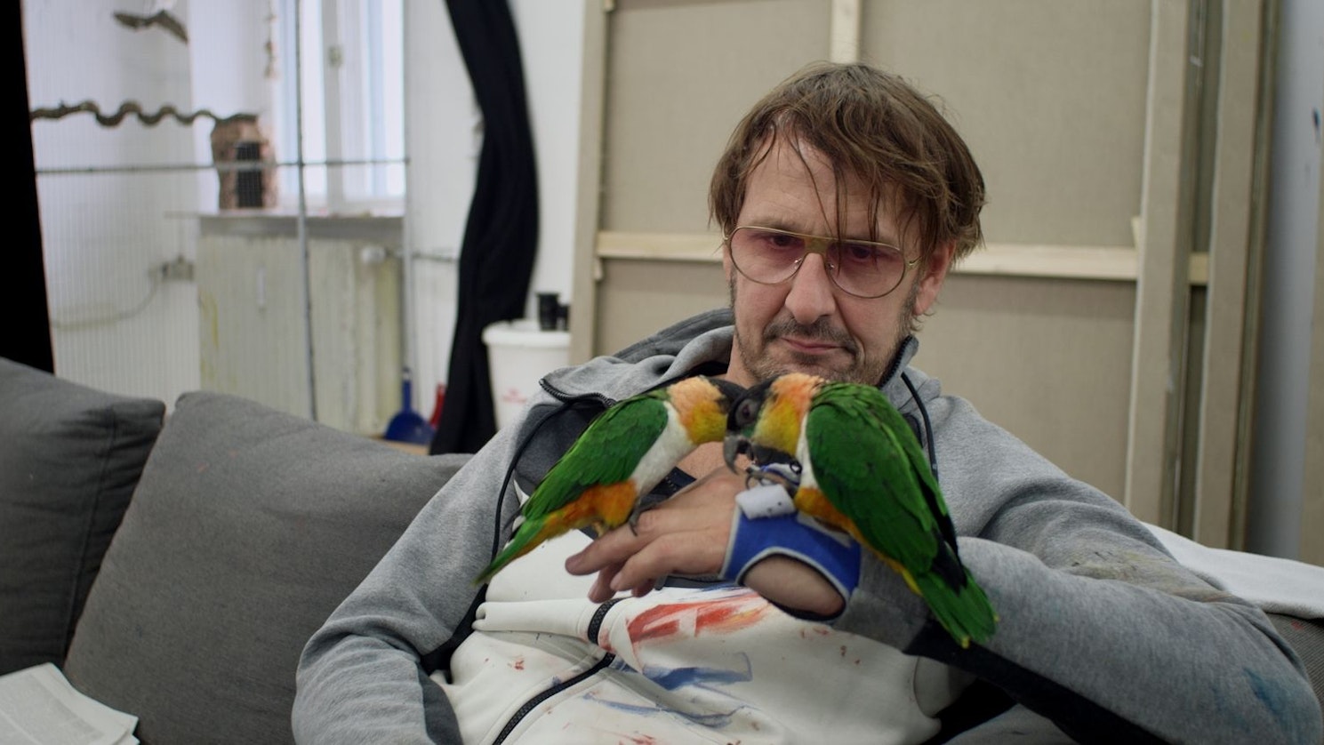 Zu sehen ist Künstler Daniel Richter, auf dessen Hand zwei Papageien sitzen.