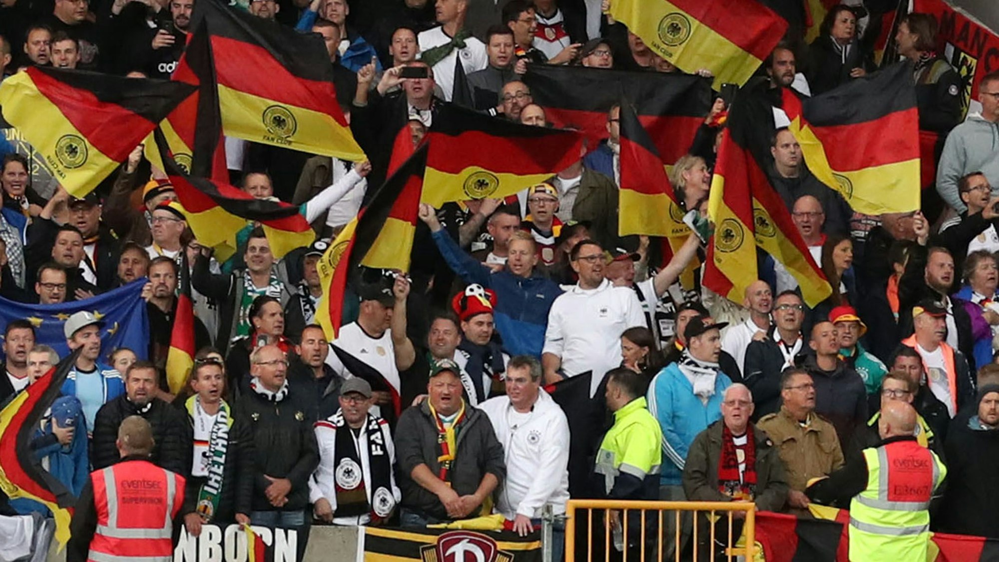 Fußball-Fans aus Deutschland stehen auf einer Tribüne und schwenken Flaggen.