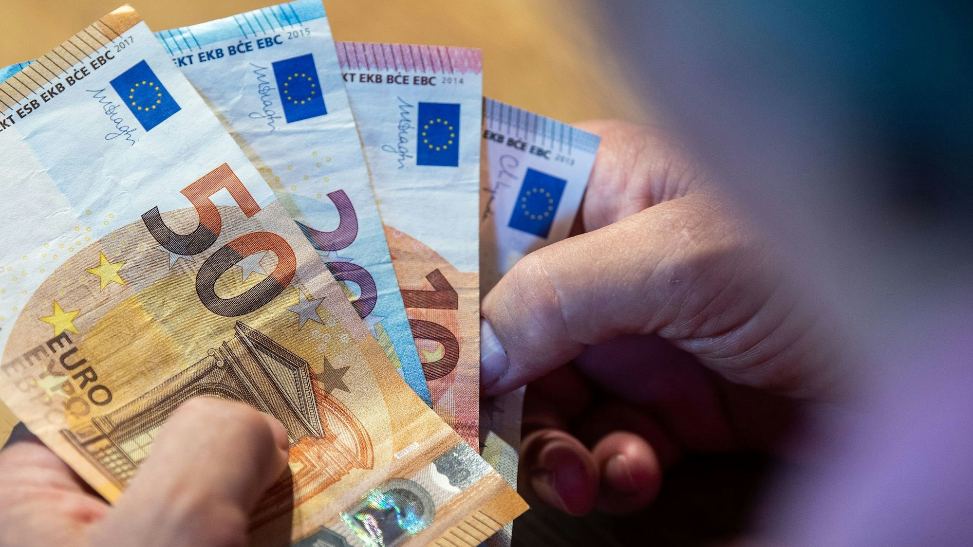 Ein älterer Mann sitzt an einem Tisch und einen 50-Euro-Schein, einen 20- und zwei 10-Euro-Scheine in der Hand.