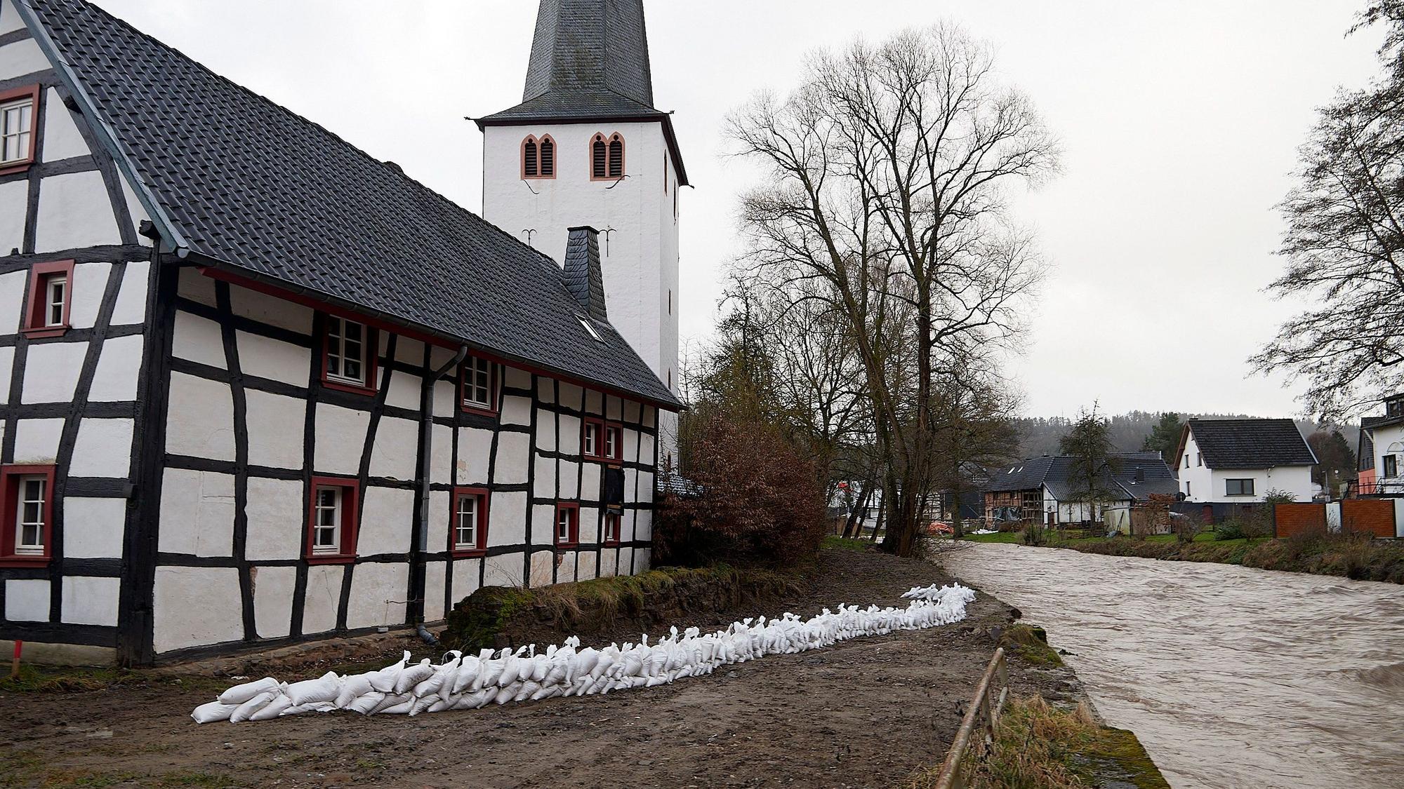 Eine Mauer aus weißen Sandsäcken ist in der Mitte des Bildes zu sehen. Rechts ist die Hochwasser führende Olef, links ein Fachwerkhaus, das durch die Säcke geschützt werden soll.