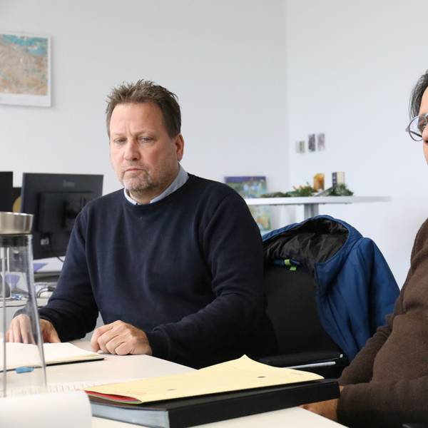 Zwei Männer sitzen an einem Tisch, auf dem Aktenmappen liegen. Im Hintergrund hängt eine mehrfarbige Stadtkarte an der Wand.