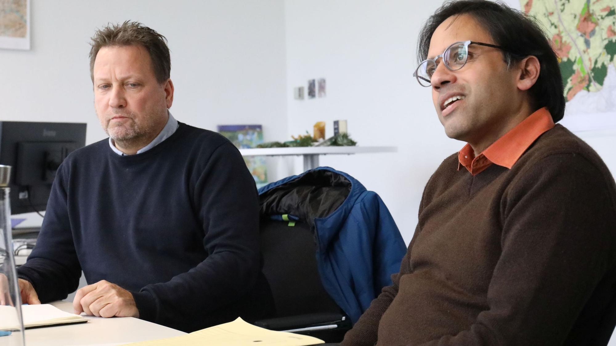 Zwei Männer sitzen an einem Tisch, auf dem Aktenmappen liegen. Im Hintergrund hängt eine mehrfarbige Stadtkarte an der Wand.
