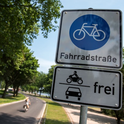 Ein Schild Fahrradstraße mit dem Zusatz Motorräder und Autos frei hängt an einer Fahrradstraße, auf der eine Radfahrerin unterwegs ist.