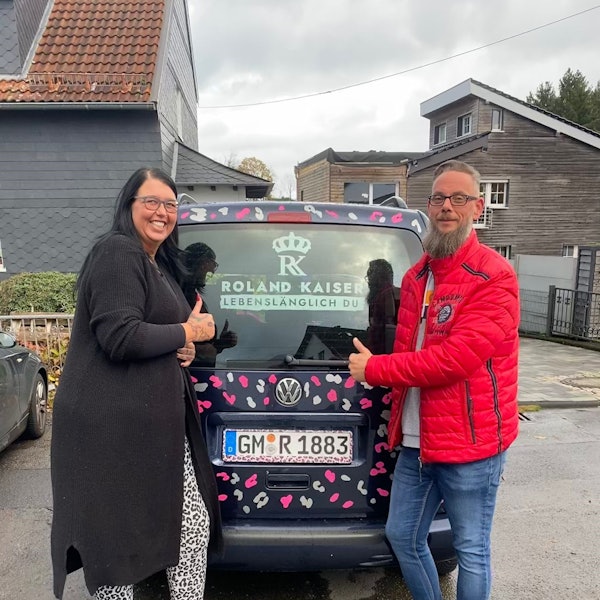 Blick auf ein Ehepaar, das hinter einem bunten Auto steht.