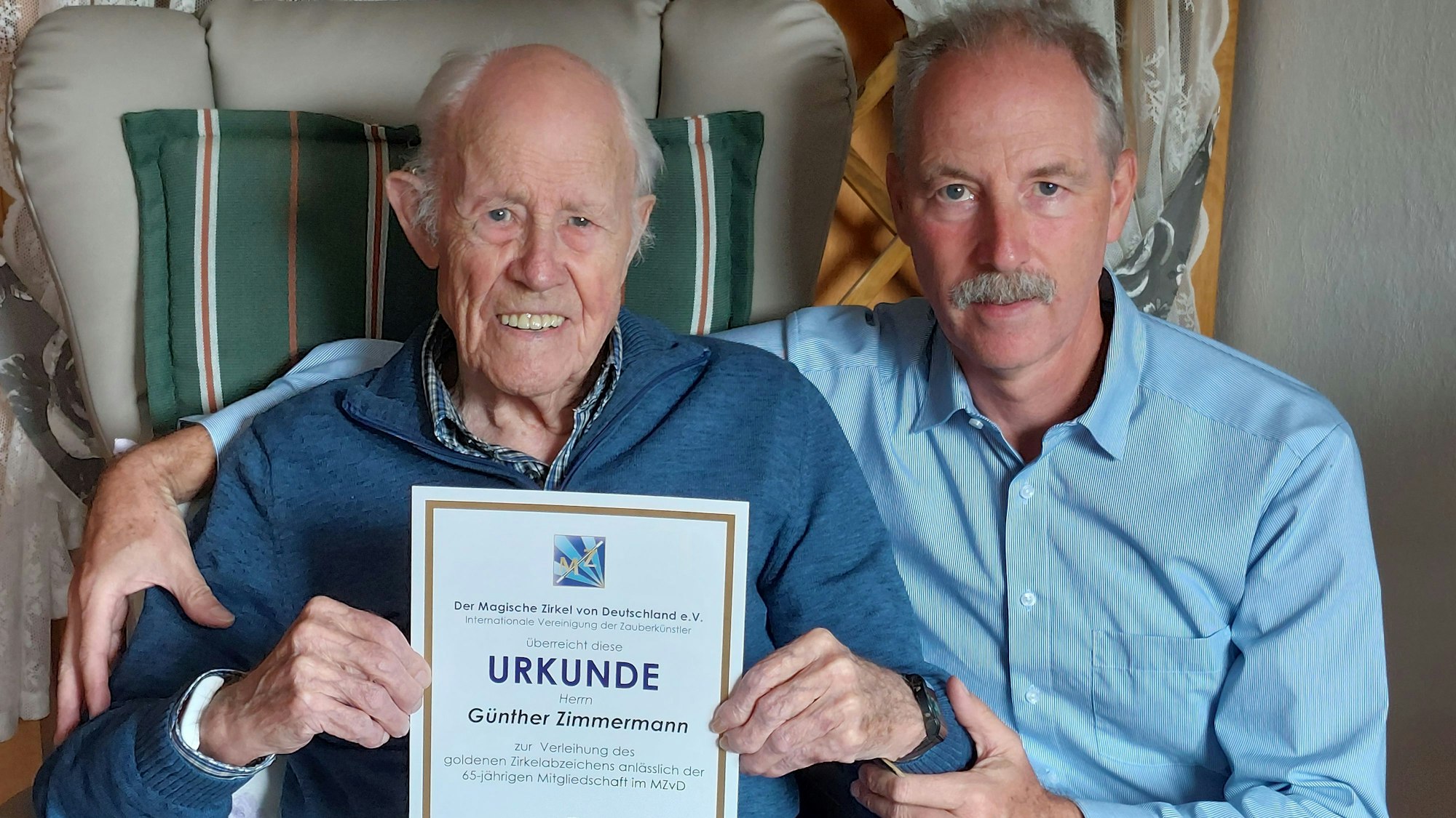 Zwei Männer sitzen nebeneinander, der linke Mann ist 96 Jahre alt und hält eine Urkunde in den Händen.