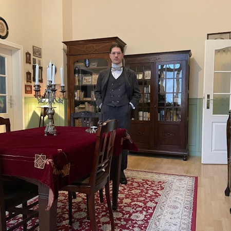 Ein Mann im altmodischen Anzug und Hemd mit Fliege steht in einem altmodisch eingerichteten Wohnzimmer.