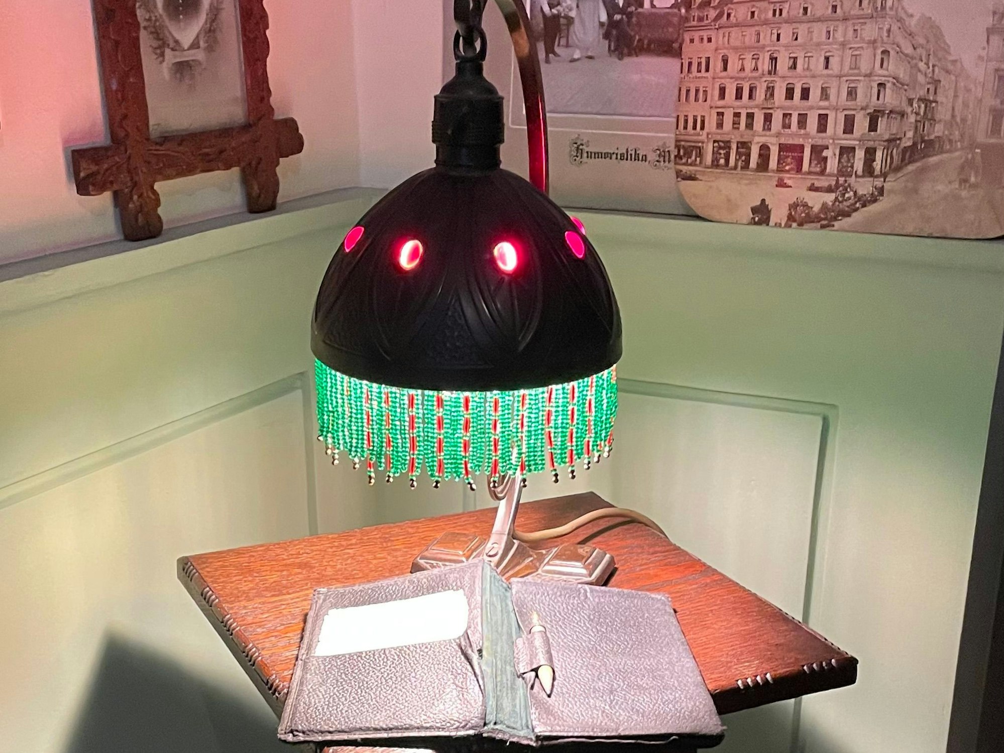 Auf einem hochbeinigen Tischchen steht eine Tischleuchte mit Fransen, an der Wand hängen alte Schwarz-Weiß-Fotos.