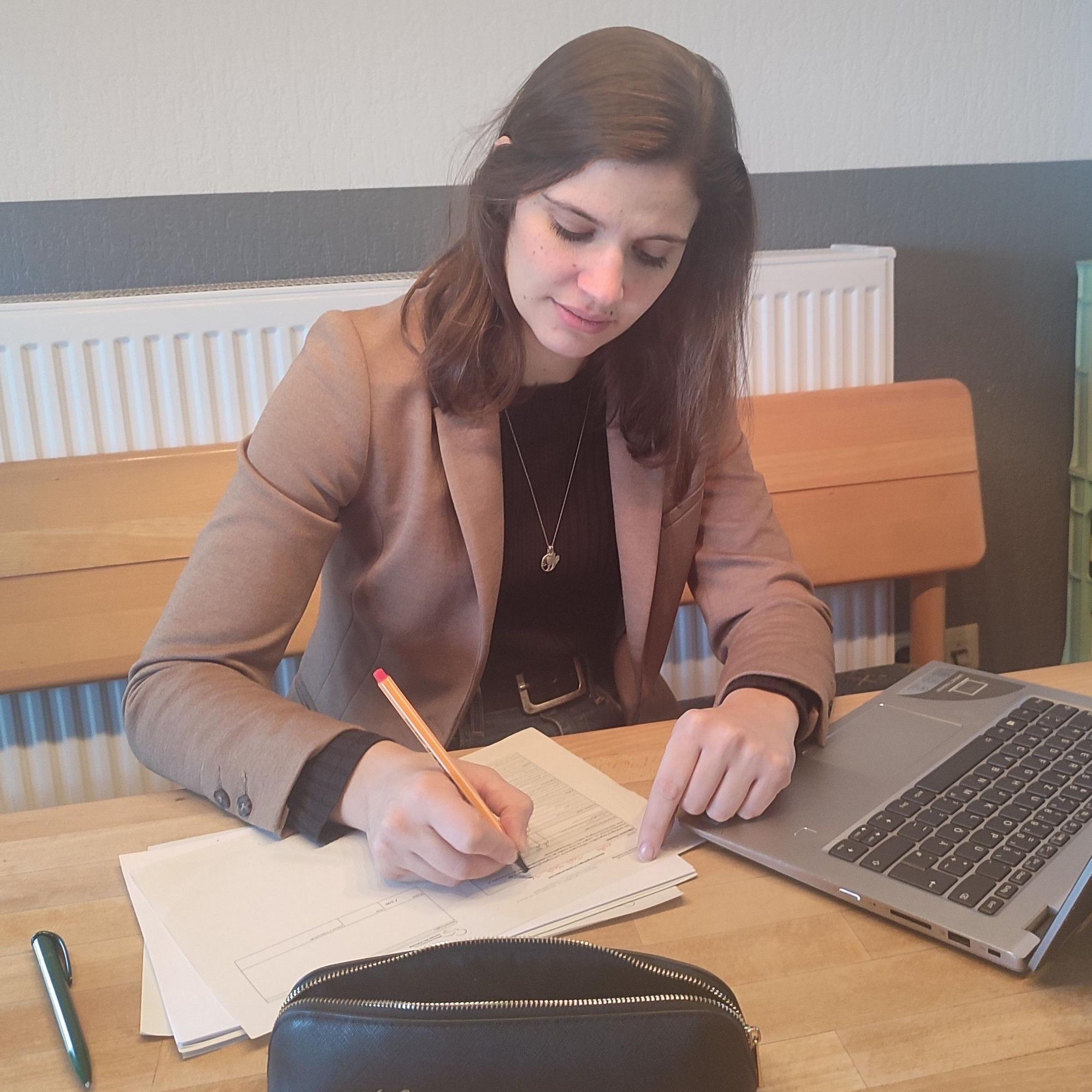 Kathrin Dahmen sitzt an einem Tisch und korrigiert mit einem Rotstift Aufsätze, vor ihr ein Laptop.