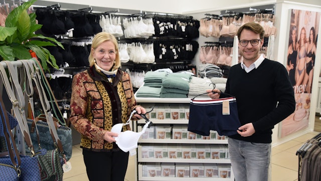 Eine ältere Frau und ein Mann stehen in einem Wäsche-Geschäft nebeneinander. Sie hält einen BH in der Hand, er Herrenunterwäsche.