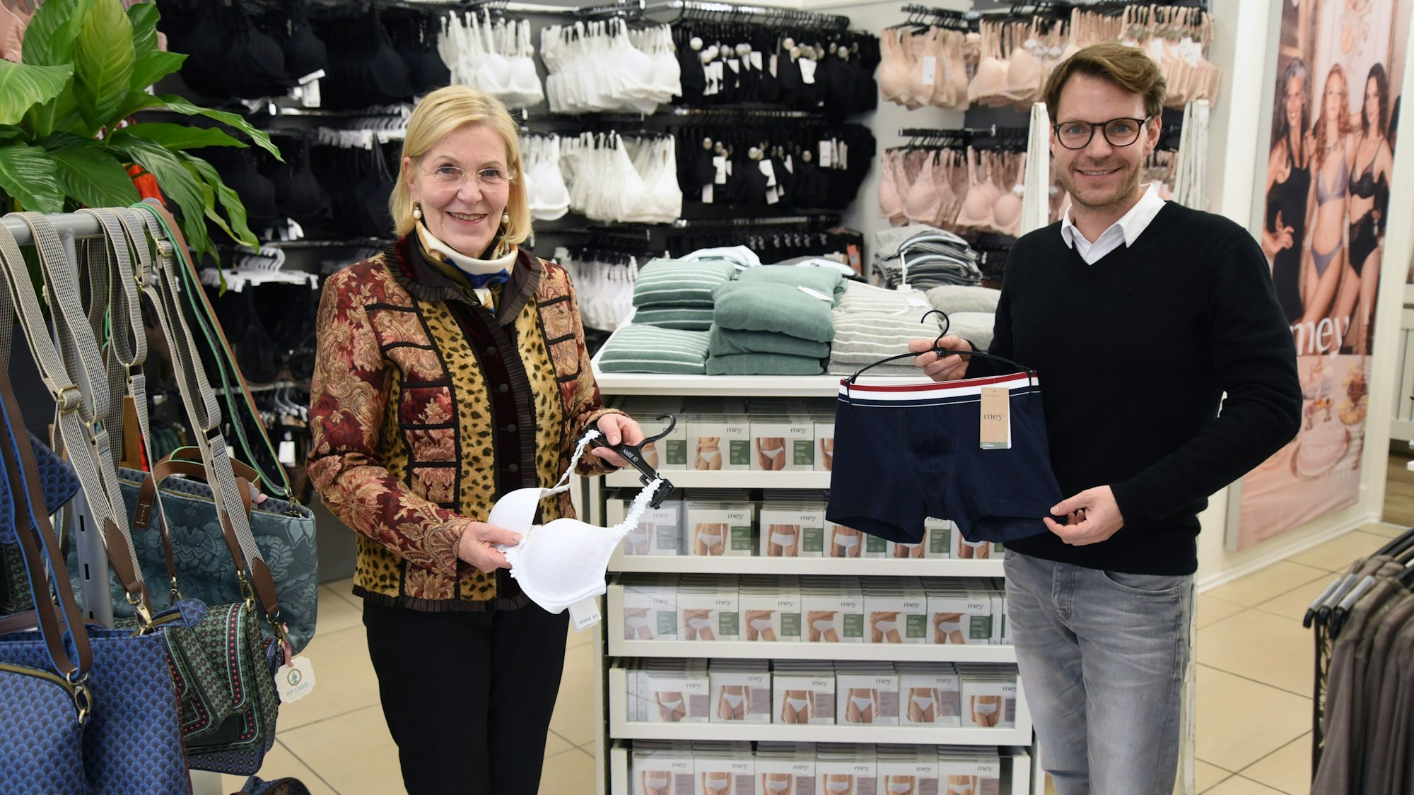 Eine ältere Frau und ein Mann stehen in einem Wäsche-Geschäft nebeneinander. Sie hält einen BH in der Hand, er Herrenunterwäsche.