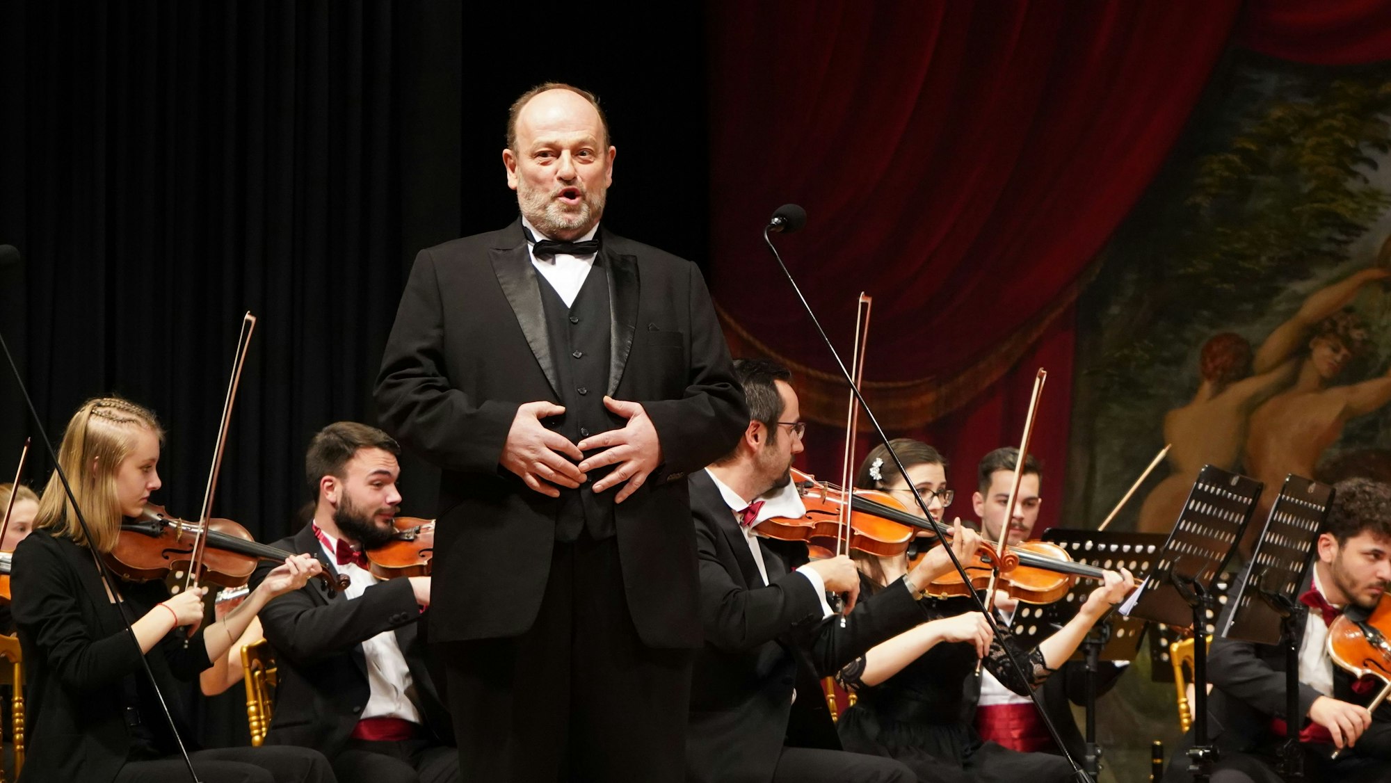 Im Vordergrund singt BaritonJosef Krenmair. Im Hintergrund spielt das Orchester.