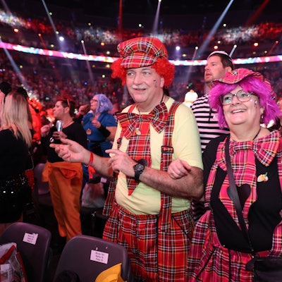 Ein verkleidetes Paar schunkelt mit eingehakten Armen, sie stehen in der Lanxess-Arena, umgeben von Feiernden.
