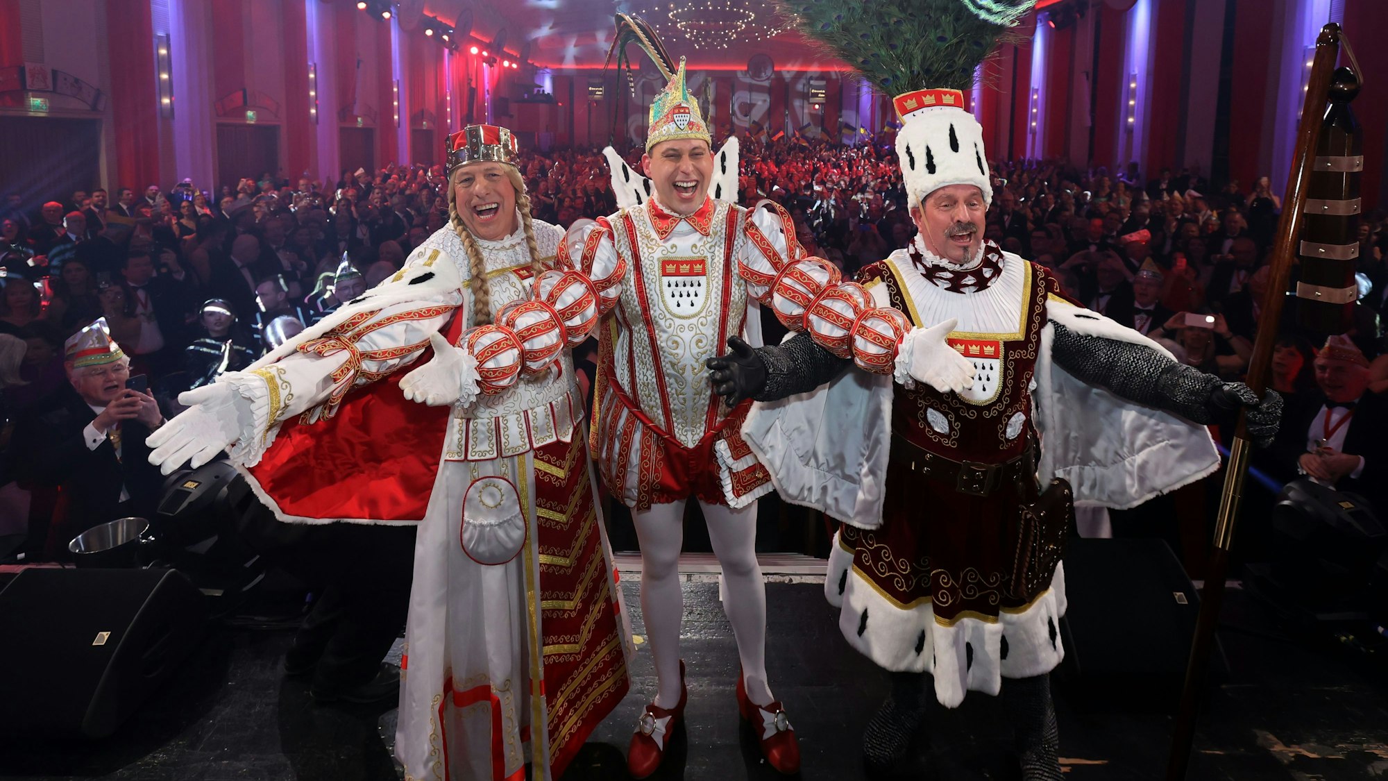 Jungfrau Frieda, Prinz Sascha I. und Bauer Werner stehen mit jeweils ausgebreiteten Armen nebeneinander auf der Bühne des Gürzenich. Sie drehen dem Publikum, das im Hintergrund zu sehen ist, den Rücken zu.
