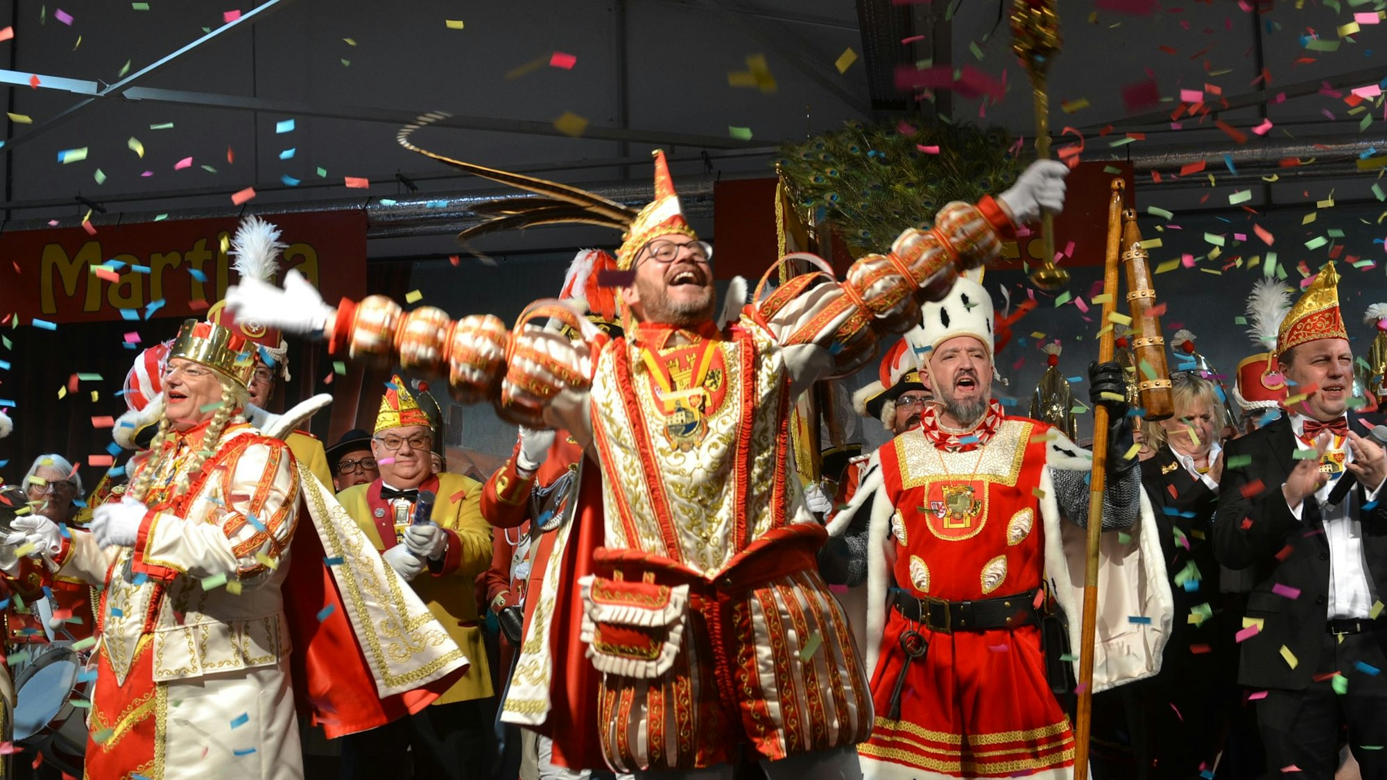 Das Dreigestirn hebt begeistert die Arme, singt und lacht. Auf die Bühne fällt ein Konfettiregen nieder.