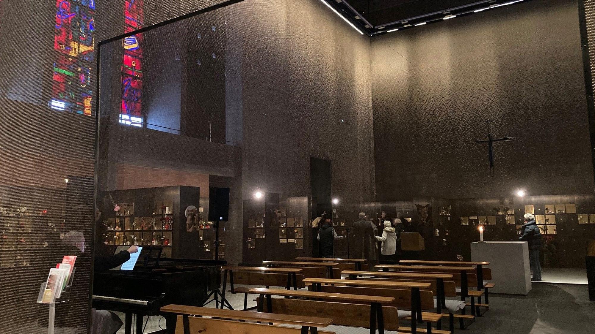 Der Innenraum einer Kirche mit Bänken, bunten Fenstern und einem filigranen netzartigen Vorhang aus Metall ist zu sehen.