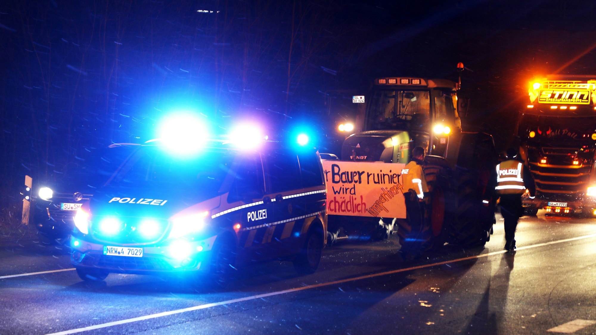 Ein Polizeiwagen mit Blaulicht steht vor einem Traktor mit Protestplakat und einem Lkw.