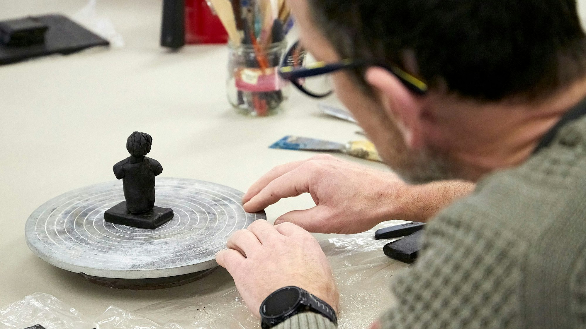 Eine kleine Skulptur aus Wachs steht auf einer drehbaren Scheibe. Ein Teilnehmer der Kunstveranstaltung schaut die Figur an, die er bearbeitet.