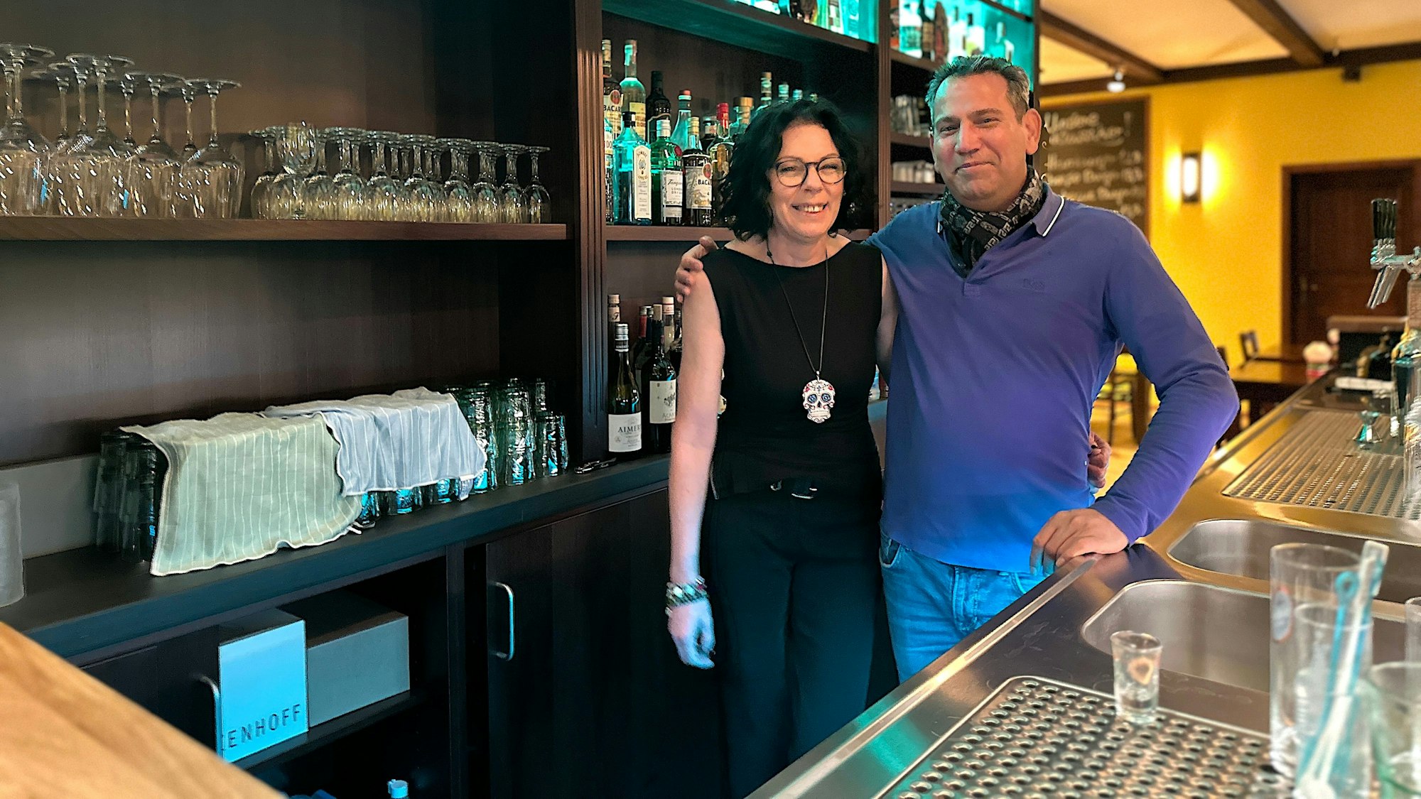 Eine dunkelhaarige Frau mit Totenkopf-Kette und ein Mann im blauen Pullover stehen hinter einer Theke.