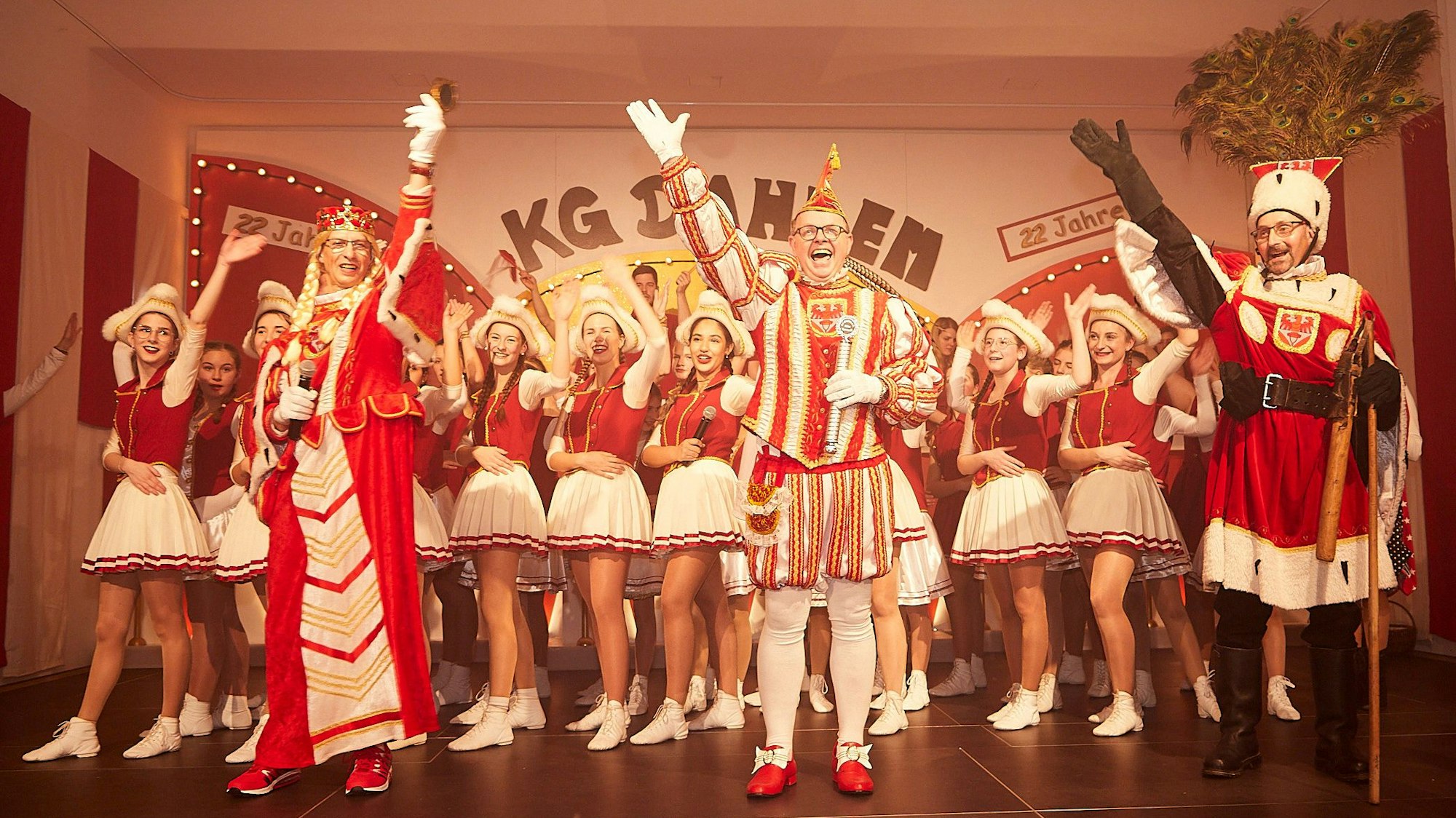 Prinz Thomas II., Bauer Thomas I. und Jungfrau Raimunde I. jubeln dem Publikum zu. Hinter ihnen auf der Bühne stehen Mitglieder einer Tanzgarde.