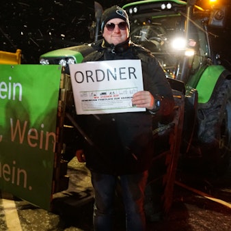 Landwirt Sebastian Bützler steht mit einem „Ordner“-Schild neben seinem Trecker auf dem Parkplatz in Breitenbenden. Auf dem Banner seines Treckers steht: „Kein Bauer, kein Bier, Schnaps, Wein – das wäre gemein!“