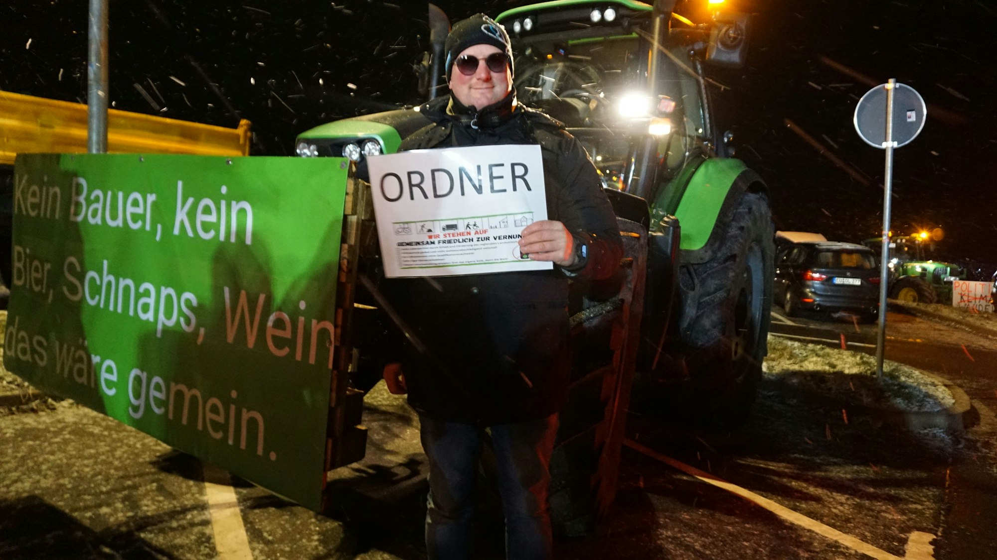 Landwirt Sebastian Bützler steht mit einem „Ordner“-Schild neben seinem Trecker auf dem Parkplatz in Breitenbenden. Auf dem Banner seines Treckers steht: „Kein Bauer, kein Bier, Schnaps, Wein – das wäre gemein!“