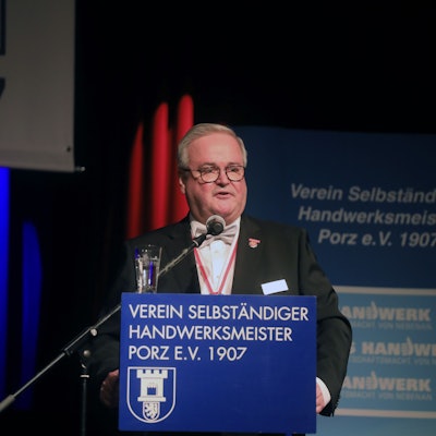 Ein Mann mit grauem Haar und Brille steht in einem Saal am Rednerpult.