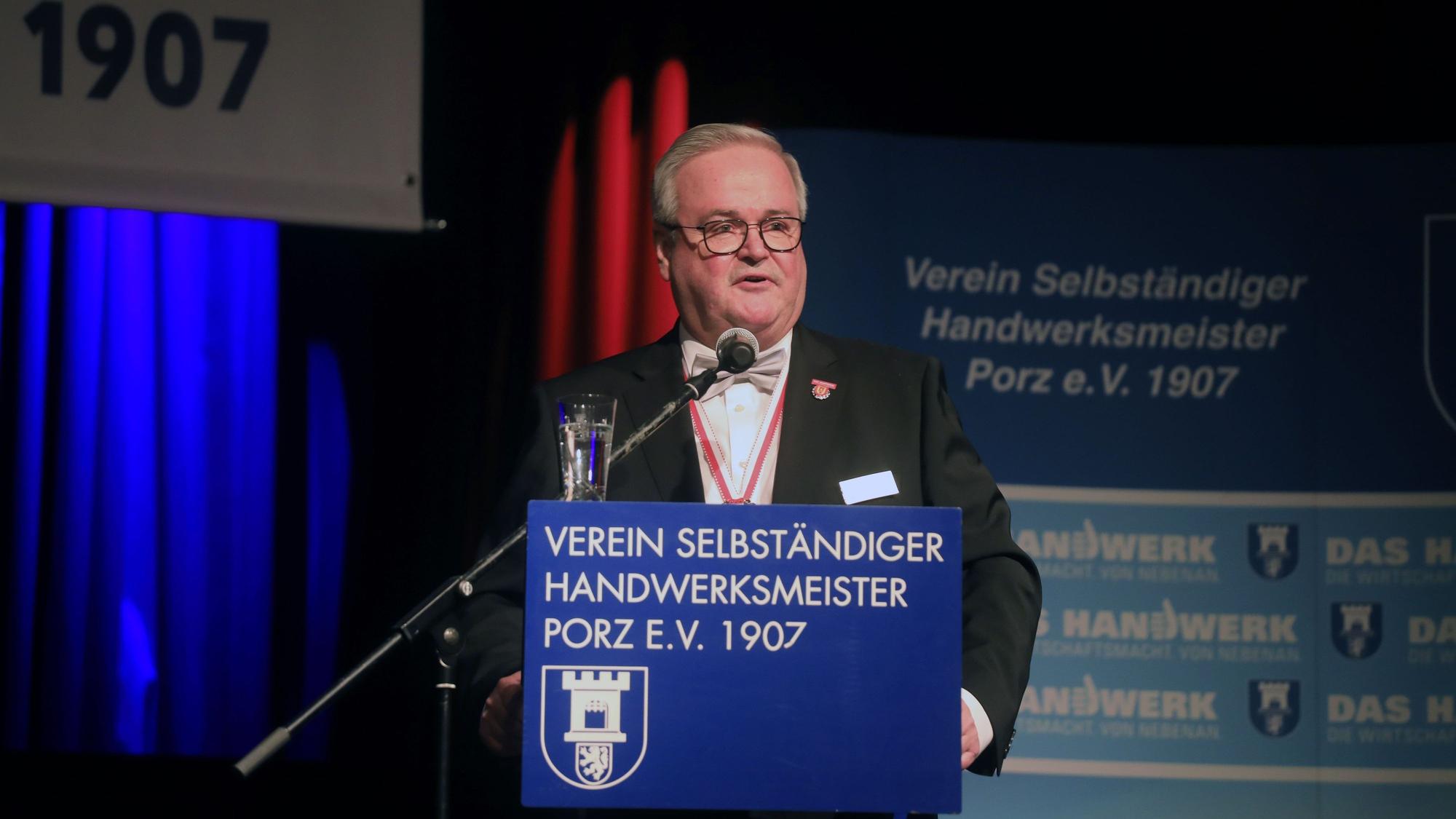 Ein Mann mit grauem Haar und Brille steht in einem Saal am Rednerpult.