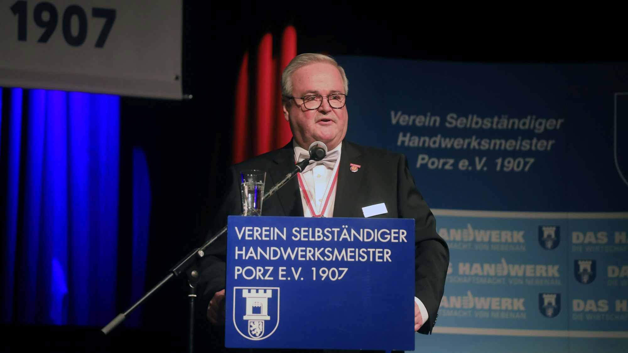 Ein Mann mit grauem Haar und Brille steht in einem Saal am Rednerpult.