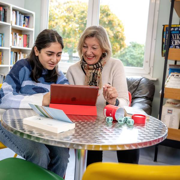 Eine Schülerin liest gemeinsam mit einer Lesementorin in der Schulbibliothek auf einem Tablet-Computer.